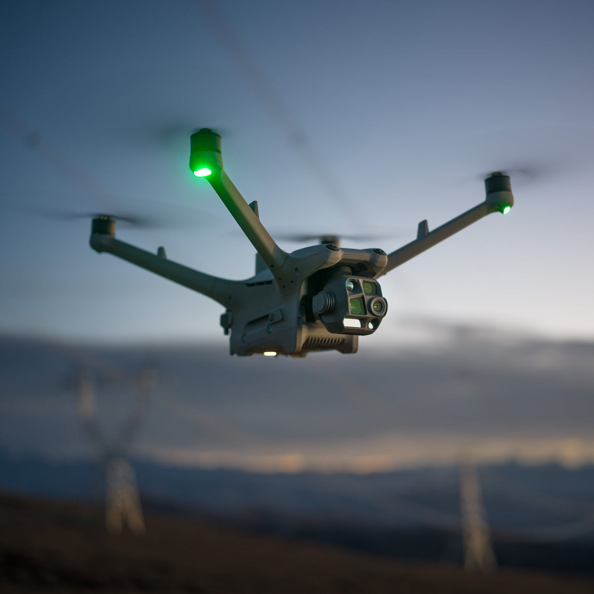 DJI Matrice 4TD in flight beside a transmission tower at dusk, the four sensor elements countable on the gimbal
